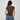 A woman viewed from behind wearing a fitted short-sleeve brown ribbed t-shirt and blue jeans, standing against a plain light gray studio background.