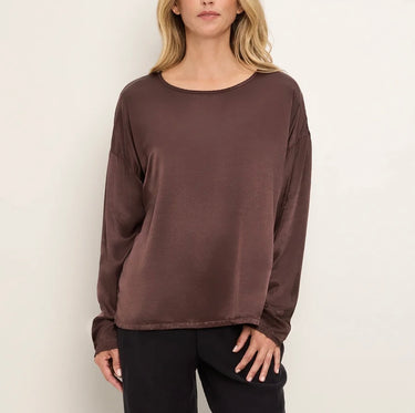 A woman wearing a long-sleeve, boat-neck top in a dark brown color, paired with black pants, standing against a plain off-white background.