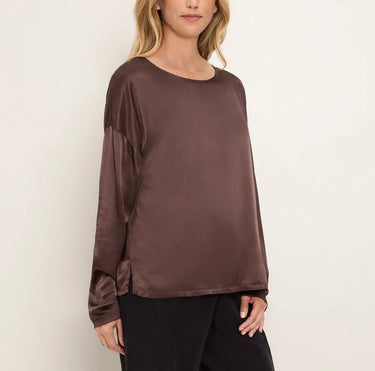 A woman wearing a long-sleeve satin blouse in a dark brown color, paired with black pants, against a plain off-white background.