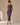 A woman modeling a sleeveless, ruched midi dress in a muted purple color, paired with clear high-heeled sandals, set against a neutral beige studio backdrop.