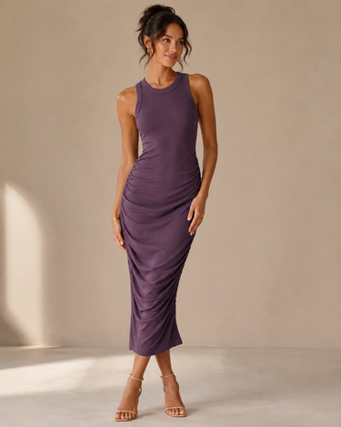 A woman modeling a sleeveless, ruched midi dress in a muted purple color, paired with clear high-heeled sandals, set against a neutral beige studio backdrop.