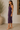 A woman modeling a sleeveless, deep purple ruched midi dress with a side slit, paired with beige block-heel mule sandals in a warm-toned indoor setting.
