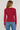 A woman viewed from the back wearing a fitted long-sleeve crewneck top in deep red and blue jeans, against a plain light gray studio background.