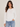 A woman modeling a white long-sleeve crewneck top with side ruching, paired with light blue jeans, against a plain light beige studio background.