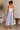 Woman wearing a white top and light blue pants, holding a beige tote bag, standing in a minimalistic room.