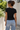 A woman viewed from behind wearing a fitted black short-sleeve t-shirt and light blue jeans, standing outdoors against a light-colored wall with greenery on the left.