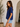 Woman wearing a blue top and denim shorts standing indoors next to a plant.
