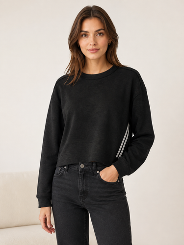 A woman modeling a black cropped crewneck sweatshirt with white stripes down the sleeves, paired with dark gray jeans, against a neutral cream background.