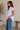 Woman wearing a light blue striped shirt and jeans standing in a room with a plant and wooden bench.
