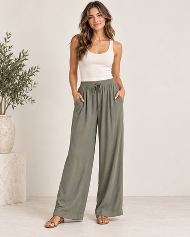 A woman modeling a pair of wide-leg, high-waisted sage green pants with a drawstring waist and side pockets, paired with a white tank top and brown sandals in a bright studio setting.