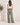 A woman shown from the back wearing wide-leg sage green pants and a white tank top, standing in a minimalist studio setting with a potted plant on the left.