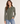 A woman modeling a long-sleeve button-up shirt in olive green, paired with blue jeans, against a plain light beige studio background.