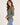 A woman wearing a long-sleeve button-up shirt in olive green, paired with light blue jeans, photographed against a plain off-white studio background.