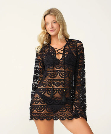 A model wearing a black bikini under a black lace long-sleeve tunic cover-up with scalloped pattern, lace-up V-neck and tassel ties.