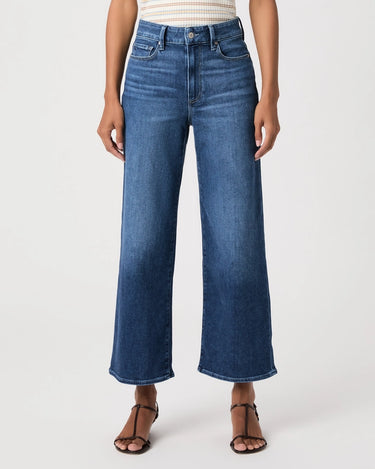 A model wearing a pair of high-waisted, wide-leg cropped blue denim jeans with a faded wash, paired with brown strappy sandals and a striped top, against a plain white studio background.