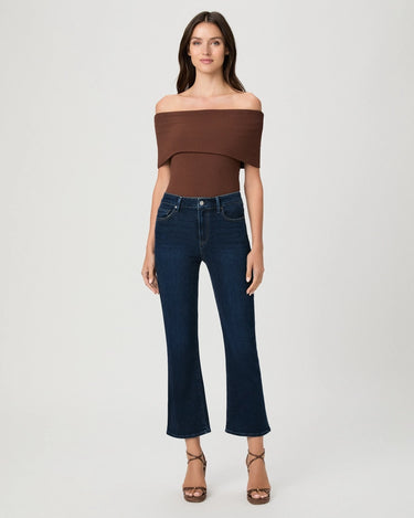 A woman modeling an off-the-shoulder brown top and dark blue cropped flare jeans with brown strappy heels, shot against a light gray studio background.