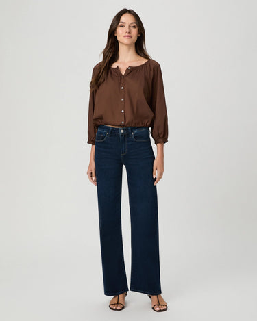 A woman modeling a brown long-sleeve button-front blouse with gathered cuffs and dark blue wide-leg jeans, standing against a light gray studio background.