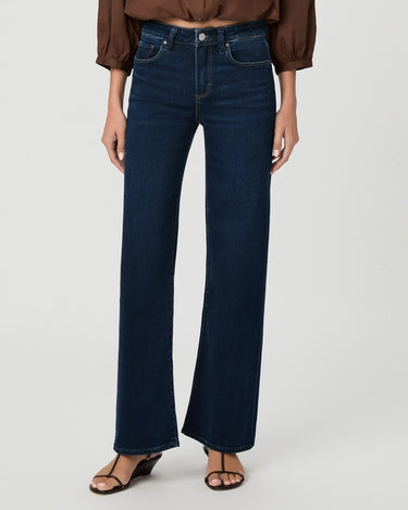 A model wearing dark blue wide-leg flare jeans, paired with a brown blouse and brown wedge sandals, shown from the waist down on a light gray studio background.