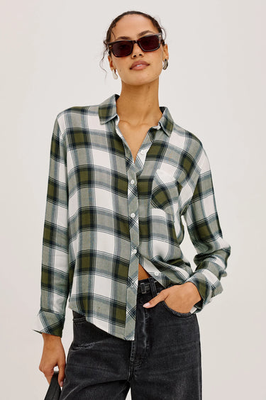A woman modeling a long-sleeve plaid button-up shirt in white, green, and gray, paired with dark jeans and sunglasses, against a plain light background.