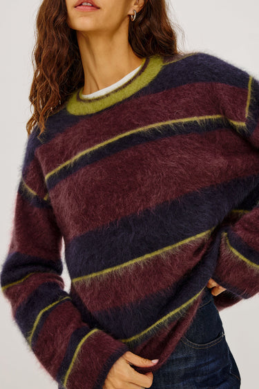 A woman wearing a burgundy, navy and green stripe brushed wool crew neck sweater and dark blue denim jeans on a white background