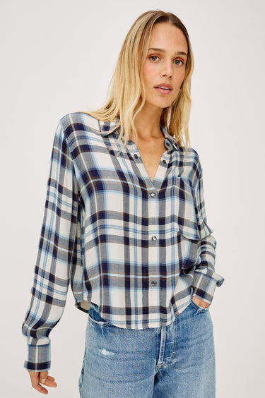 A woman wearing a long-sleeve button-up plaid shirt in blue, white, and navy, paired with light-wash blue jeans, photographed against a light gray studio background.