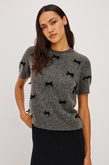 A woman wearing a short sleeve charcoal sweater with velvet black bows and a black midi skirt on a white background.