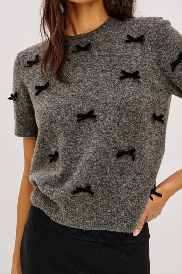 A woman wearing a short sleeve charcoal sweater with velvet black bows and a black midi skirt on a white background.