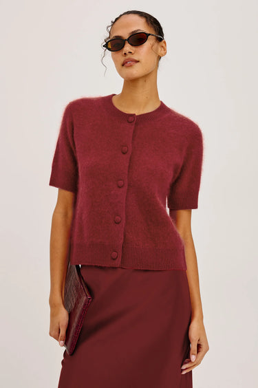 A model wearing a short-sleeve button-front knit cardigan in a deep burgundy red, paired with a matching skirt and sunglasses, against a plain light gray studio background.