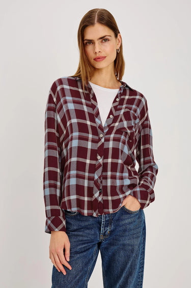 A woman modeling a long-sleeve button-front plaid shirt in burgundy, gray, and white, paired with blue jeans against a light gray studio background.