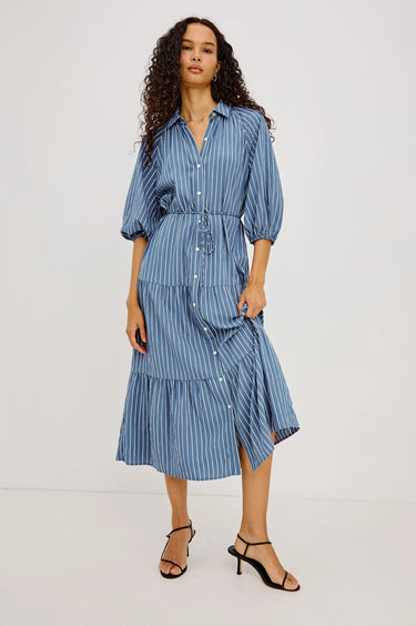 A woman modeling a blue and white vertically striped midi shirt dress with puff sleeves, a button-down front, and a tiered skirt, paired with black strappy heels against a plain white studio background.