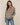 A woman modeling a long-sleeve button-up plaid shirt in cream, brown, and black, paired with blue jeans, against a plain beige studio background.