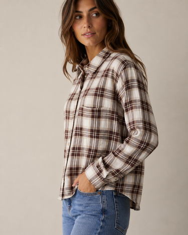 A woman wearing a long-sleeve plaid button-up shirt in cream, brown, and beige tones, paired with blue jeans, photographed against a plain light beige studio background.