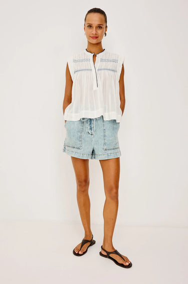 Woman wearing a white sleeveless top and light blue denim shorts on a plain background
