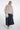 A model wearing a light beige faux fur cropped jacket with a high collar and buttons, paired with a dark navy blue leather skirt and holding a matching beige faux fur handbag against a plain white studio background.