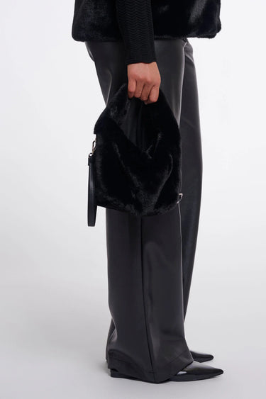A person wearing black wide-leg leather pants, a black fuzzy top, and black pointed shoes holds a black fuzzy handbag with a small black pouch attached, photographed against a light gray studio background.
