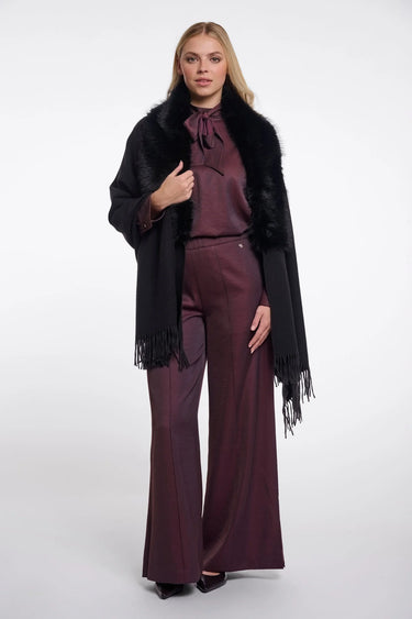 A woman modeling a black fringed faux-fur cape over a matching dark plum satin top and wide-leg trousers, photographed against a light gray studio background.