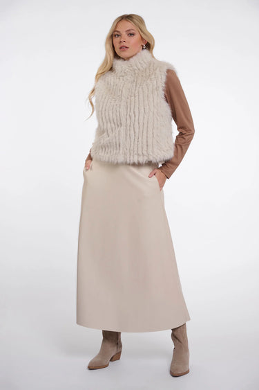 A woman modeling a sleeveless cream faux-fur vest over a brown long-sleeve top, paired with a beige midi skirt and taupe ankle boots, against a light gray studio background.