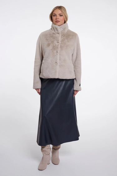 A model stands against a light gray studio background wearing a light beige cropped faux-fur jacket with a high collar and three front buttons, paired with a dark navy midi skirt and beige ankle boots.