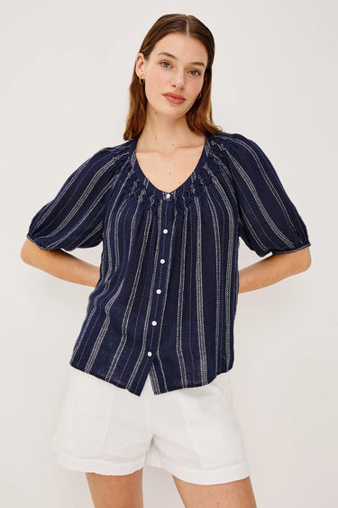 Woman wearing a navy striped blouse with white buttons and white shorts on a light background