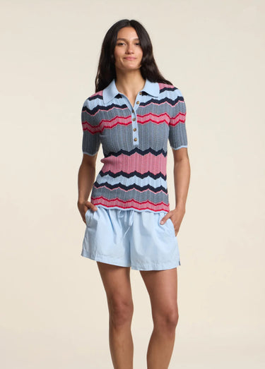 A woman wearing a zig zag stripe polo in multi color with blue shorts on a neutral background