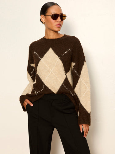 A woman wearing a brown and cream argyle knit sweater with black trousers, styled with gold-framed sunglasses, standing against a plain off-white wall.