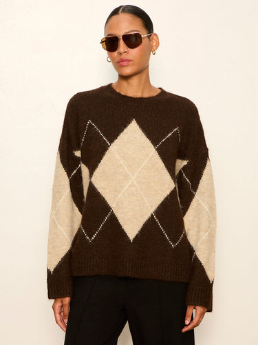 A woman wearing a dark brown and beige argyle knit sweater, styled with dark sunglasses and black pants, standing against a plain off-white wall.