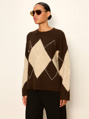A woman wearing a brown and cream argyle knit sweater and black pants, standing against a plain off-white wall.
