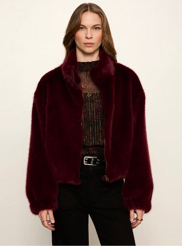 A woman modeling a plush, cropped fur jacket in deep burgundy. The jacket features a wide, high collar and is styled over a dark patterned top and black pants against a plain off-white studio background.
