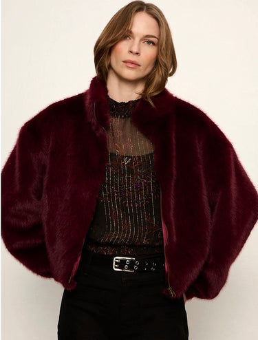 A woman wearing a plush, cropped burgundy faux fur jacket over a sheer black top and black pants, photographed against a plain off-white background.