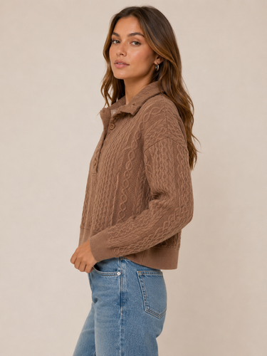 A woman wearing a brown cable-knit cropped sweater with a buttoned collar, paired with light blue jeans, standing against a plain beige studio background.