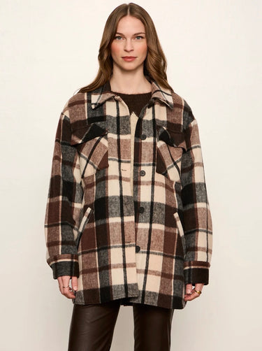 A woman modeling a brown, beige, and black plaid button-up coat over a dark brown top and dark brown leather pants, set against a plain off-white studio background.