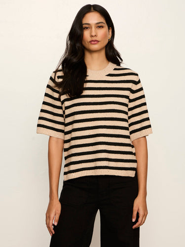 A woman modeling a short-sleeve knit sweater with horizontal beige and black stripes, paired with black pants, against a plain off-white background.