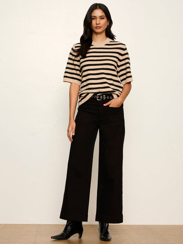 A woman modeling a beige and black horizontally striped short-sleeve knit top with black wide-leg pants and black ankle boots, standing against a plain off-white wall.