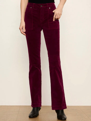A person wearing deep burgundy corduroy pants with a slight flare at the hem, paired with black ankle boots, standing against a plain off-white wall.